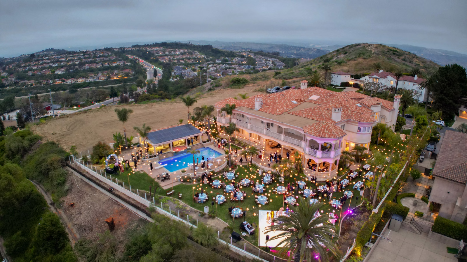 An elevated view of a hillside estate, pool and dining lawn illuminated for an evening celebration.
