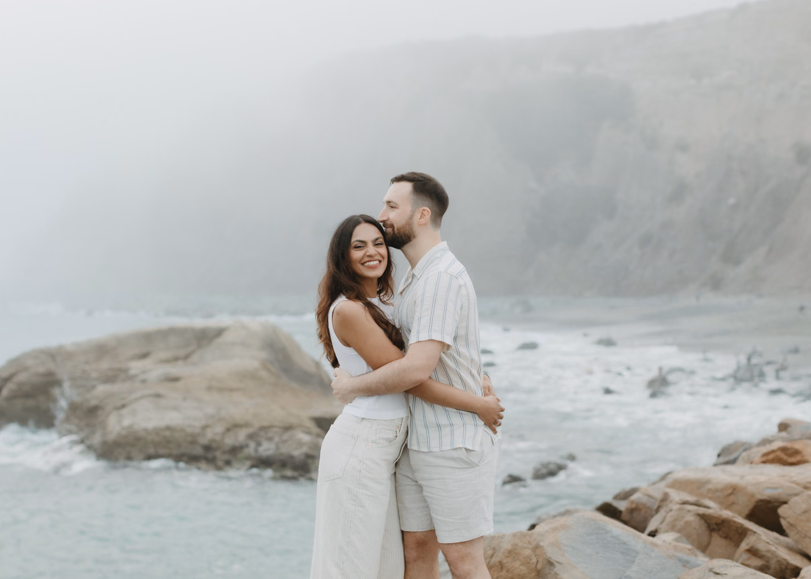 Anu smiling as John kisses her temple during a quiet embrace on the coast