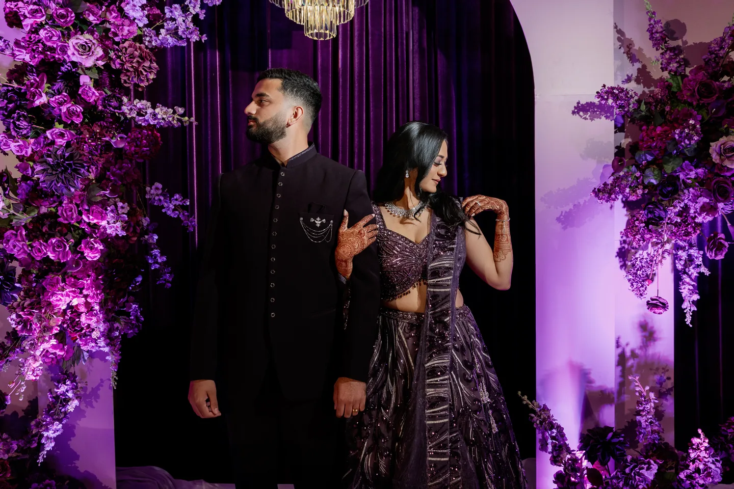 Mahima and Sarang framed by purple reception florals, chandeliers and draped staging