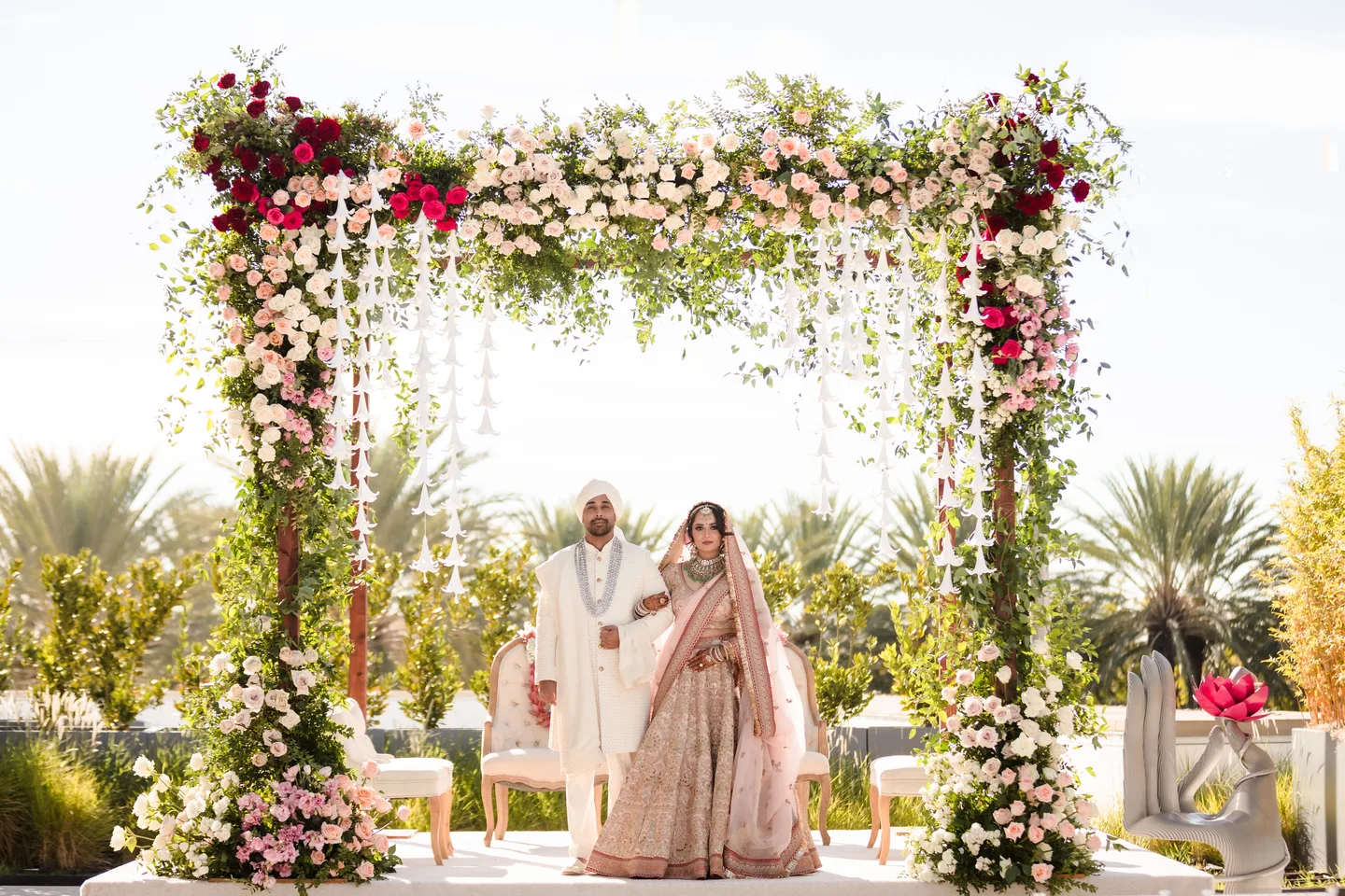 Natasha and Anish together beneath a complete floral ceremony structure, framed by palms and garden surroundings