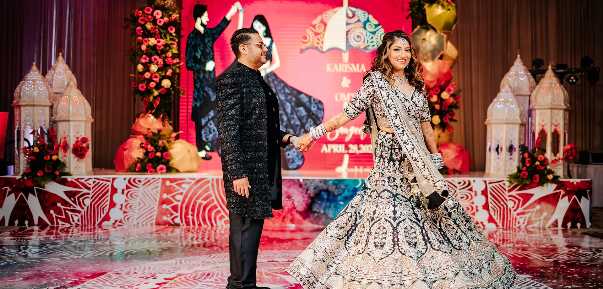 Karisma and Om in coordinated Sangeet attire against a richly colored floral stage.