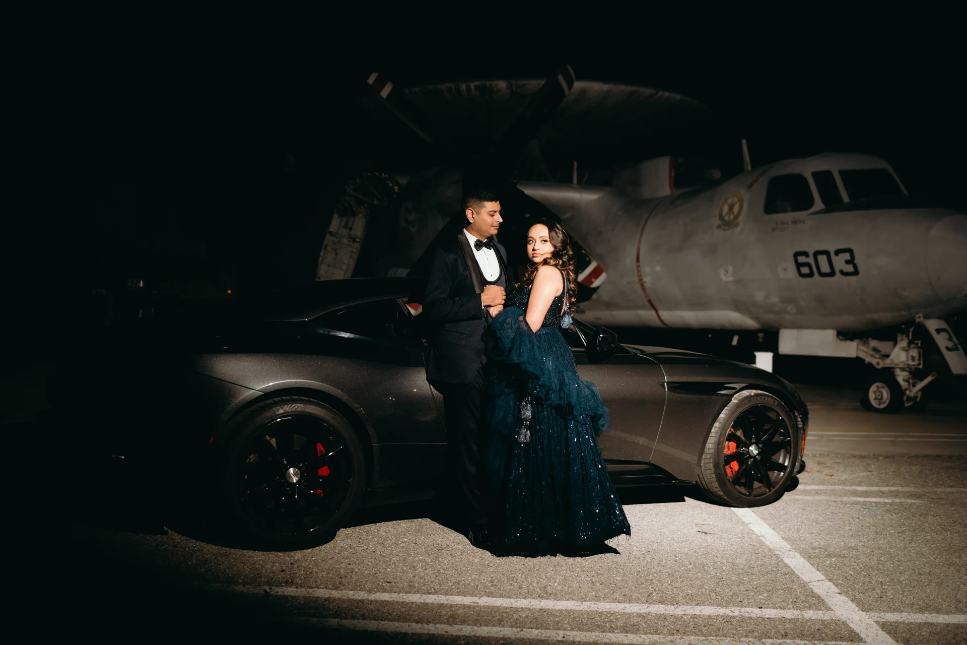 Purna and Mayur together in evening attire beside a sports car and aircraft